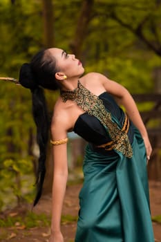 An Indonesian woman poses with black hair dangling to the ground while wearing a green dance costume inside the forest