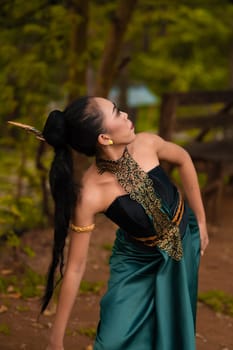 An Indonesian woman poses with black hair dangling to the ground while wearing a green dance costume inside the forest