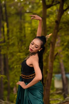 A brave Asian woman in makeup wearing a green costume and gold jewelry with the woods in the background during the morning