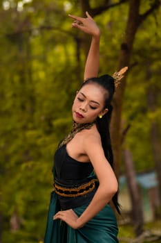 A brave Asian woman in makeup wearing a green costume and gold jewelry with the woods in the background during the morning