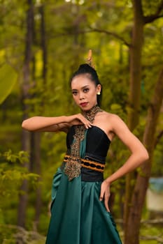 A brave Asian woman in makeup wearing a green costume and gold jewelry with the woods in the background during the morning