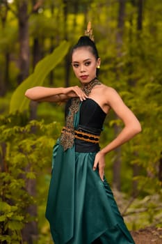A brave Asian woman in makeup wearing a green costume and gold jewelry with the woods in the background during the morning
