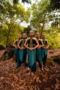 a wide shot of an Asian woman standing together in the forest while wearing a green dance costume with dead leaves in the background