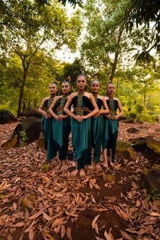 a wide shot of an Asian woman standing together in the forest while wearing a green dance costume with dead leaves in the background