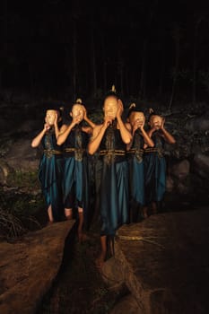 A Group of Asian women wearing a wooden masks in green costumes while standing between the rocks inside the forest during the night