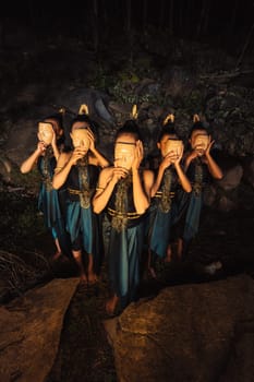 A Group of Asian women wearing a wooden masks in green costumes while standing between the rocks inside the forest during the night