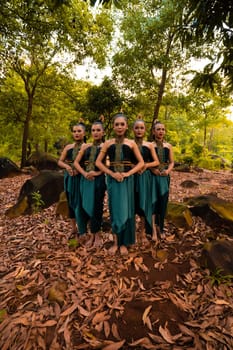 a wide shot of an Asian woman standing together in the forest while wearing a green dance costume with dead leaves in the background