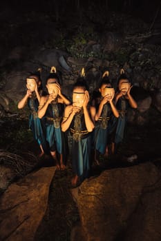 A Group of Asian women wearing a wooden masks in green costumes while standing between the rocks inside the forest during the night