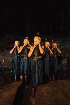 A Group of Asian women wearing a wooden masks in green costumes while standing between the rocks inside the forest during the night