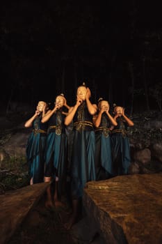 A Group of Indonesian women holding and wearing a brown wooden mask in a green skirt between the rock inside the woods