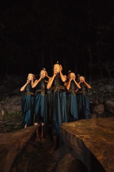 A Group of Indonesian women holding and wearing a brown wooden mask in a green skirt between the rock inside the woods