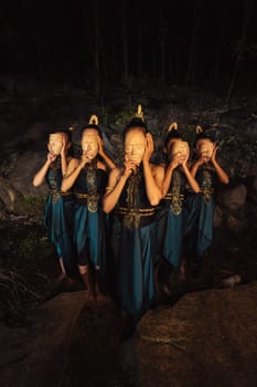 A Group of Asian women wearing a wooden masks in green costumes while standing between the rocks inside the forest during the night