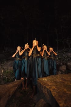 A Group of Indonesian women holding and wearing a brown wooden mask in a green skirt between the rock inside the woods