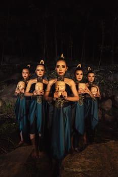 A Bunch of girls in green costumes holding a wooden masks in their hands while standing between the big rocks in front of the forest