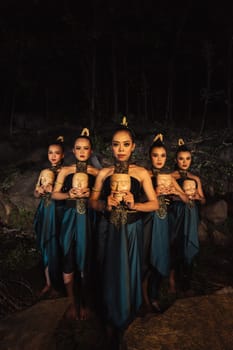 A Bunch of girls in green costumes holding a wooden masks in their hands while standing between the big rocks in front of the forest