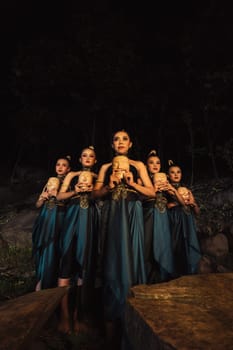 A Bunch of girls in green costumes holding a wooden masks in their hands while standing between the big rocks in front of the forest