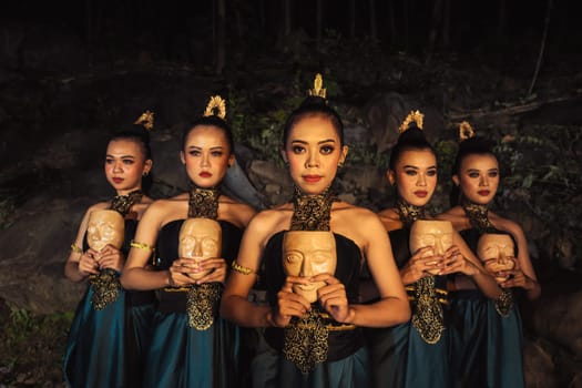 A Bunch of girls in green costumes holding a wooden masks in their hands while standing between the big rocks in front of the forest