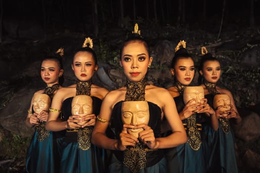 A Group of ethnical women holding a wooden craft mask in their hands while in line inside the wood in the night