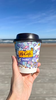 Miami, FL USA - February 05, 2025: A hand holds a McCafe coffee cup featuring playful doodles against a stunning blue sky and sandy beach, highlighting the joyful experience of enjoying a beverage