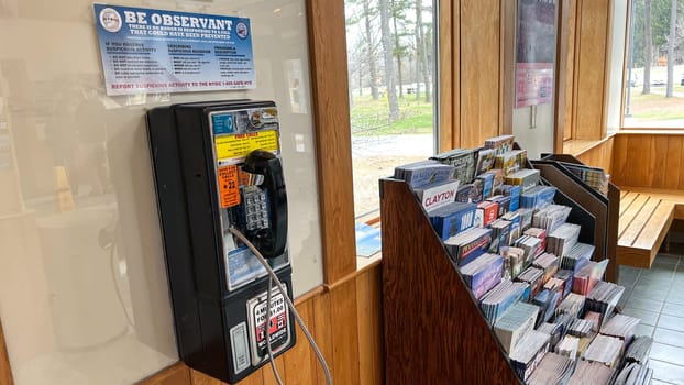Syracuse, NY USA - March 04, 2025: A vintage payphone is mounted on a wooden wall next to a brochure rack filled with colorful travel pamphlets in a public space illuminated by natural light
