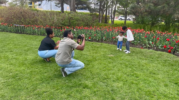 Ottawa, Canada - May 09, 2025: African American family enjoying a sunny day in a park, capturing joyful moments with a camera, Tulip festival and lush green grass, showcasing love and togetherness.
