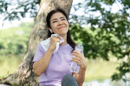 A content woman relaxes by the lake, using a towel to wipe sweat, reflecting wellness and active lifestyle.