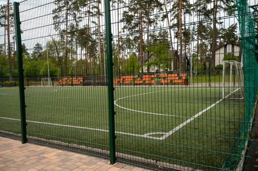 football field with white lines on the grass and orange stands. Tall trees around. surrounded by a green net, a fence around the football field in the form of an iron net with large cells.