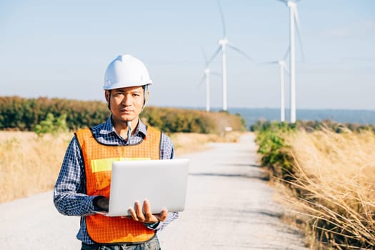 Engineer with laptop near wind turbines. A service technician ensures efficiency using innovation for landscape development. Working confidently ensuring quality in windmill service.
