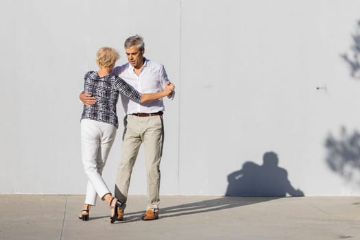 Happy retired couple dancing tango in the street in front of a white wall