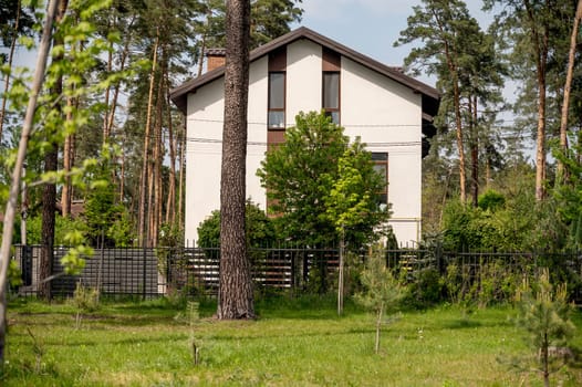 Beautiful exterior of new house with facade and brown roof two-storey house. White residential houses in the village surrounded by pine forest. Various trees around the house.