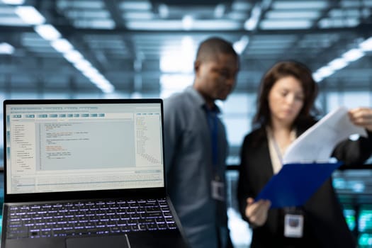 Close up of code lines on laptop screen in front of data center employees reading paperwork. Focus on programming language on notebook display next to server hub workers reading technical files