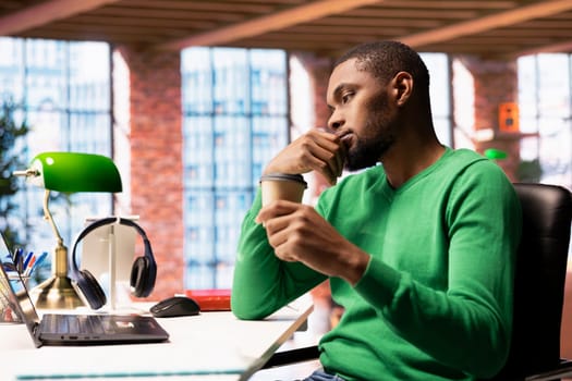 Man holding coffee cup enjoying watching TV show during relaxation time at home. Person sitting at desk entertained by engrossing television series on laptop, enjoying beverage