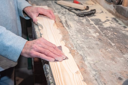 Close-up of carpenter's hands skillfully sanding decorative wooden plank on workbench in carpentry workshop, surrounded by various tools and small sawdust