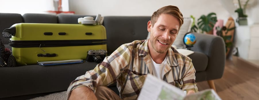Portrait of handsome smiling man, holding map, sitting with suitcase in living room, going abroad on holiday, studying his route. Tourism concept