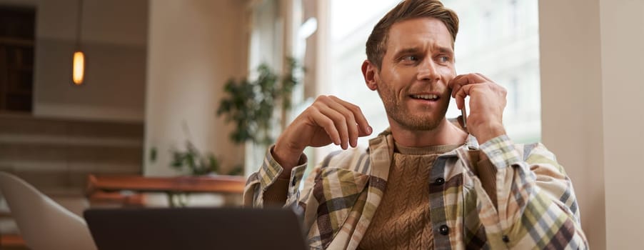 Portrait of young businessman, freelancer working remotely from coffee shop in city, sitting in chair with laptop, answer a phone call, talking on mobile to someone.