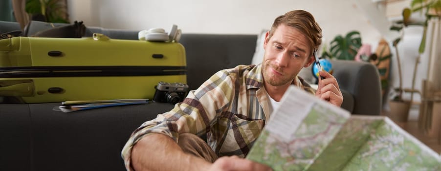 Portrait of man, tourist looking confused at travel map, shrugging, cant decide on route, choosing the road for travelling. Tourism concept