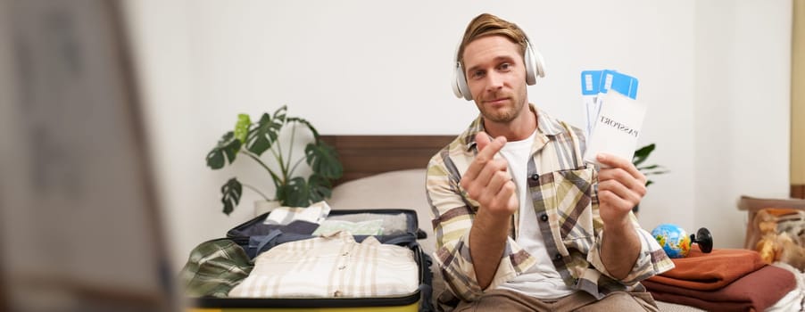 Happy tourist, young man shows passport and tickets, goes on holiday, sits with packed suitcase on bed, plans summer vacation.