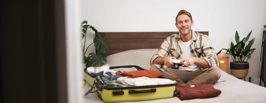 Portrait of young excited man, sits on bed with suitcase, going on holiday, holding digital camera, going through photo album on gadget, preparing for summer vacation.