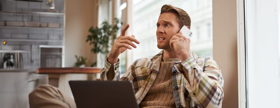 Image of man feeling stressed while talking over the phone. Businessman sitting in cafe with laptop, answering a call, hear bad news over the telephone.