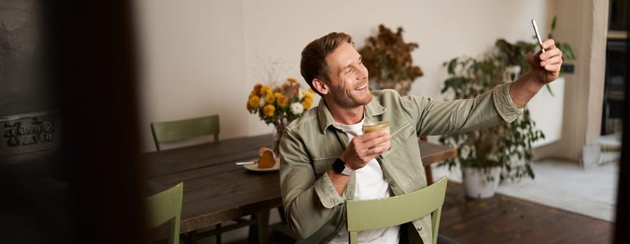Portrait of handsome happy young man, taking selfie in cafe, posing with cup of coffee, video chats using smartphone app, talks to a friend. Lifestyle and leisure concept