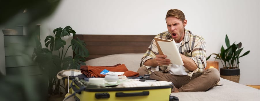 Portrait of angry man, packing suitcase, looking frustrated at notebook with check list of items for holiday, preparing to go on vacation. Tourist and people concept