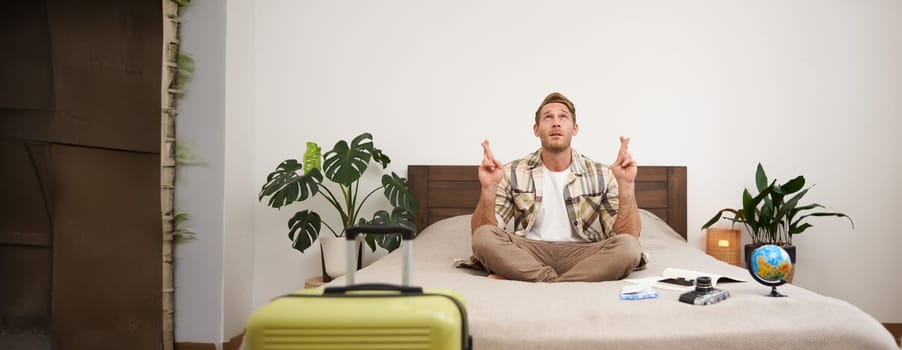 Portrait of young man meditating with fingers crossed, praying, making a wish, packing suitcase to go on holiday, has globe and camera on a bed.