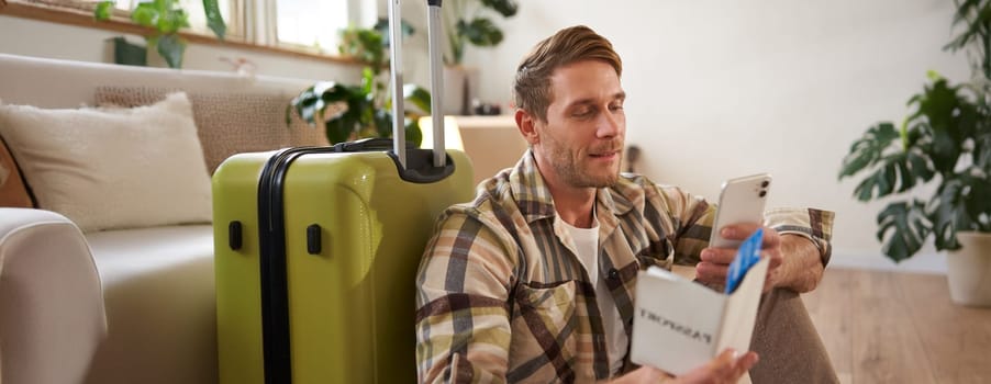 Close up portrait of handsome young man, sits on suitcase, holds passport, flight tickets and his mobile phone, using smartphone app to check departure time.