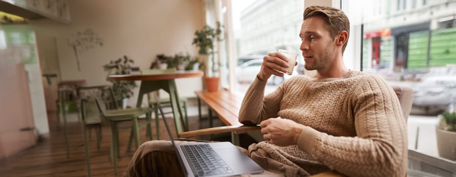 Working people and public space concept. Young handsome man, ux ui designer sitting with laptop in cafe, drinking cappuccino, looking at screen with serious face.