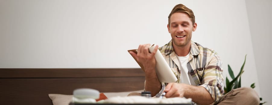 Portrait of tourist packing suitcase, going through check-list, writing down items to take with him in luggage, making notes in notebook, sitting on bed.