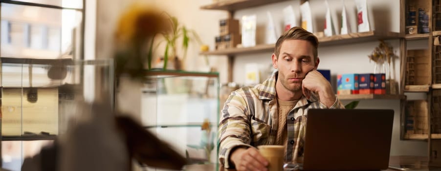 Portrait of man looking sad and stressed at his glass of coffee, working from cafe, sitting with laptop, lacks inspiration, doesnt feel inspired to finish the project.