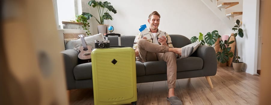 Portrait of man sits with mobile phone in living room, packed suitcase, chats on smartphone app, looks for holiday destinations, goes on vacation.