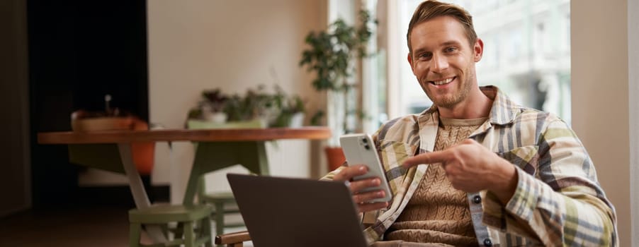 Portrait of young man freelancer, working from cafe, sitting on chair with laptop, pointing finger at his phone screen, showing interest in something on his phone.