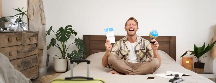 Portrait of cheerful, happy tourist, man sitting with plane tickets and a globe, packed suitcase for travelling around the world, going on vacation, excited about his holiday.