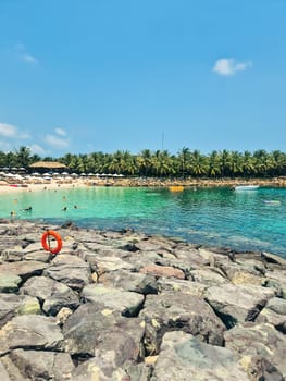 Vertical beach shot in Vietnam showing rocky shoreline with a lifebuoy in foreground. Crystal clear water, distant swimmers, palm trees and horizon